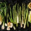 An overhead view of asparagus, lettuce, green onions, garlic, and a lemon cooking on a grill over charcoal.