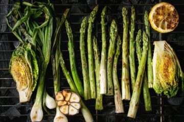 An overhead view of asparagus, lettuce, green onions, garlic, and a lemon cooking on a grill over charcoal.
