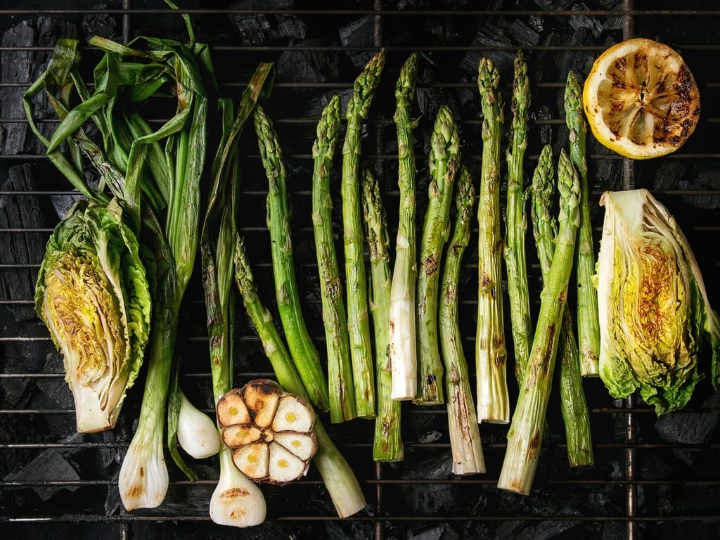 An overhead view of asparagus, lettuce, green onions, garlic, and a lemon cooking on a grill over charcoal.