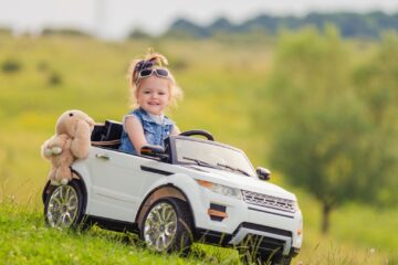 A little girl wearing white sunglasses and a denim vest is driving a white electric ride-on car with a stuffed bunny.