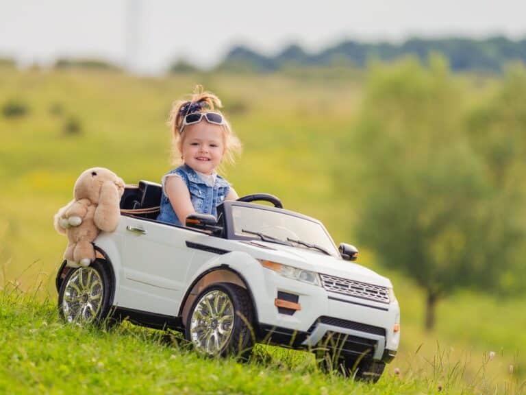 A little girl wearing white sunglasses and a denim vest is driving a white electric ride-on car with a stuffed bunny.