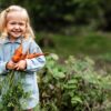 A young blonde girl wearing a denim shirt stands in a garden smiling, holding a handful of freshly picked carrots.