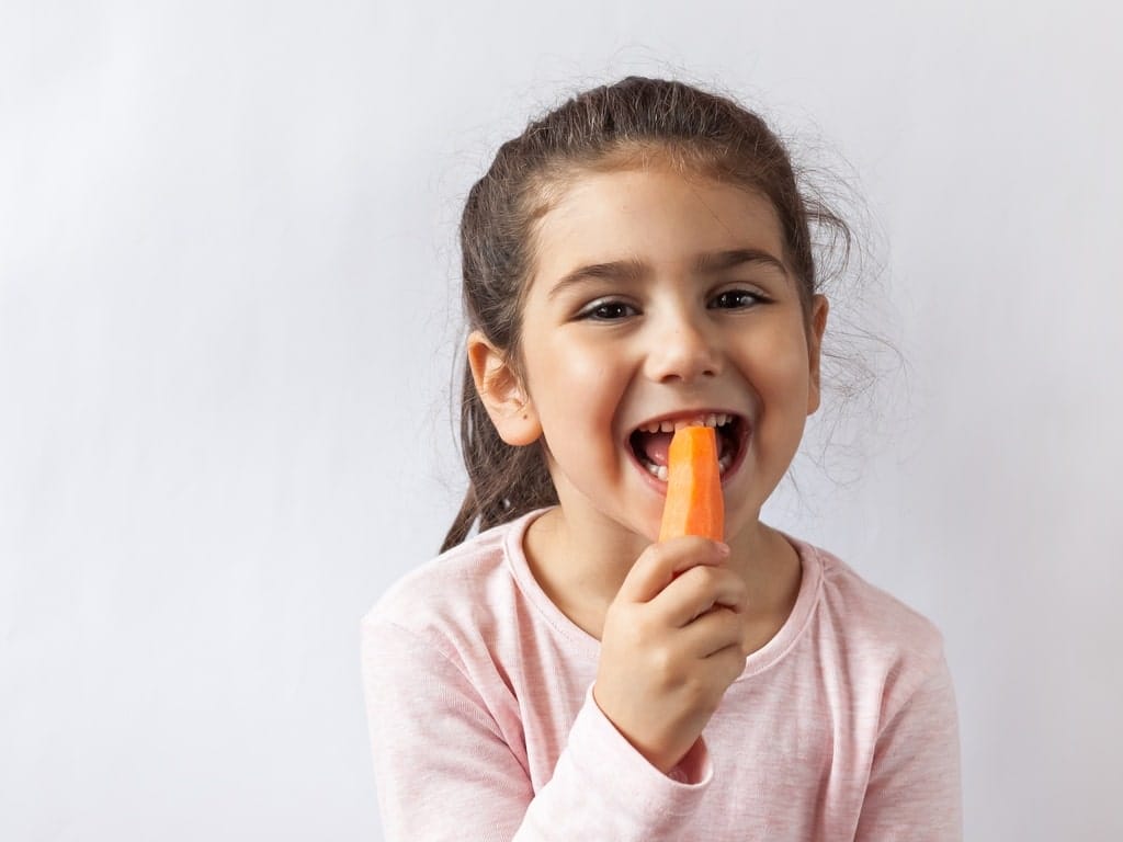 A young girl in a pink shirt and a ponytail smiling while eating a carrot with the skin peeled off.