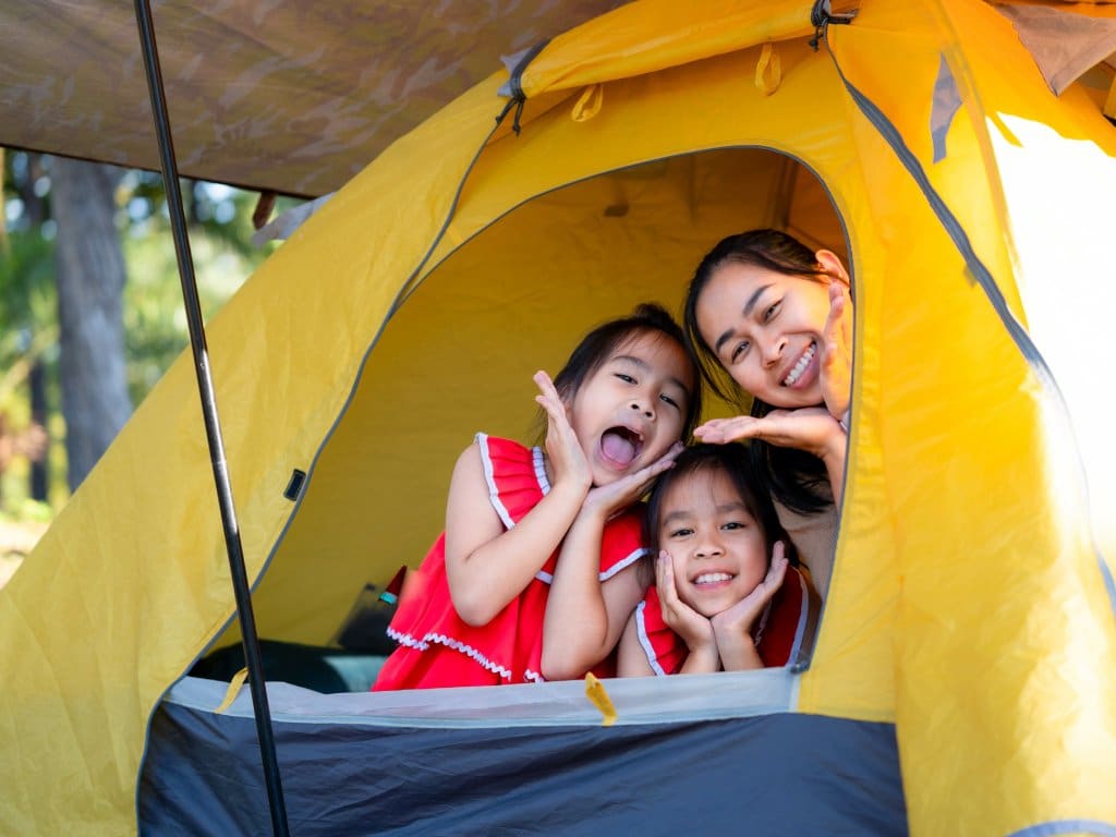 A mom and her two daughters place their faces in the opening of a yellow tent, making funny faces and smiling.