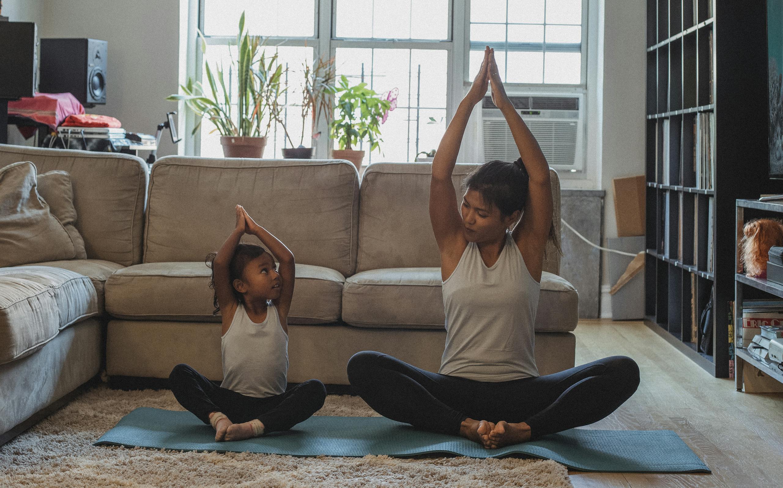 Full body content fit Asian woman and daughter in activewear sitting in Lotus Pose with arms raised and looking at each other while practicing yoga in cozy living room