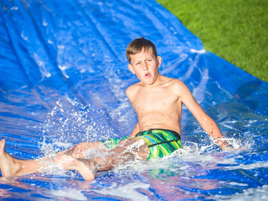 A blue tarp is on top of a grassy hill with water on it. A boy sits at the bottom of the tarp with water splashing around him.