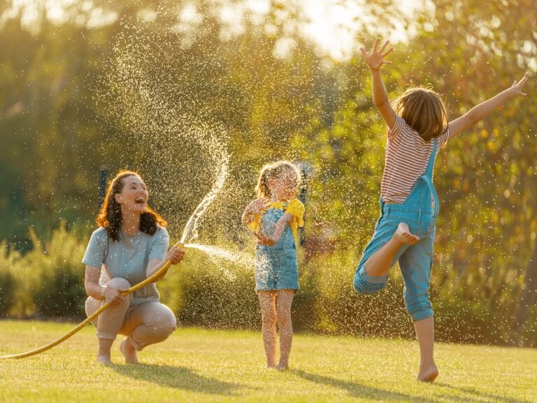 A smiling woman spraying a garden hose in a grassy area while two blonde children play in the water.