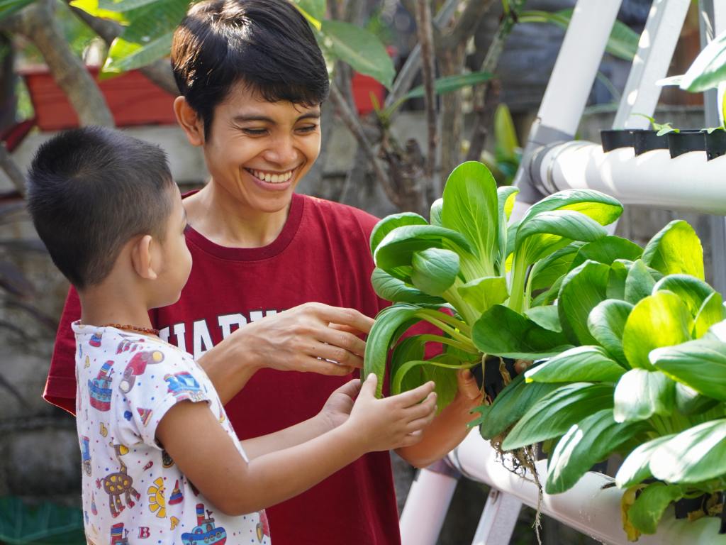 A mother and son smile as they harvest multiple small vegetables from a white hydroponic system.