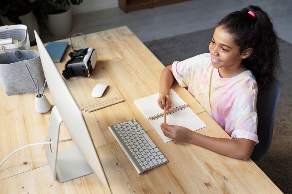 A young girl smiling while studying online at home, using a computer and taking notes.