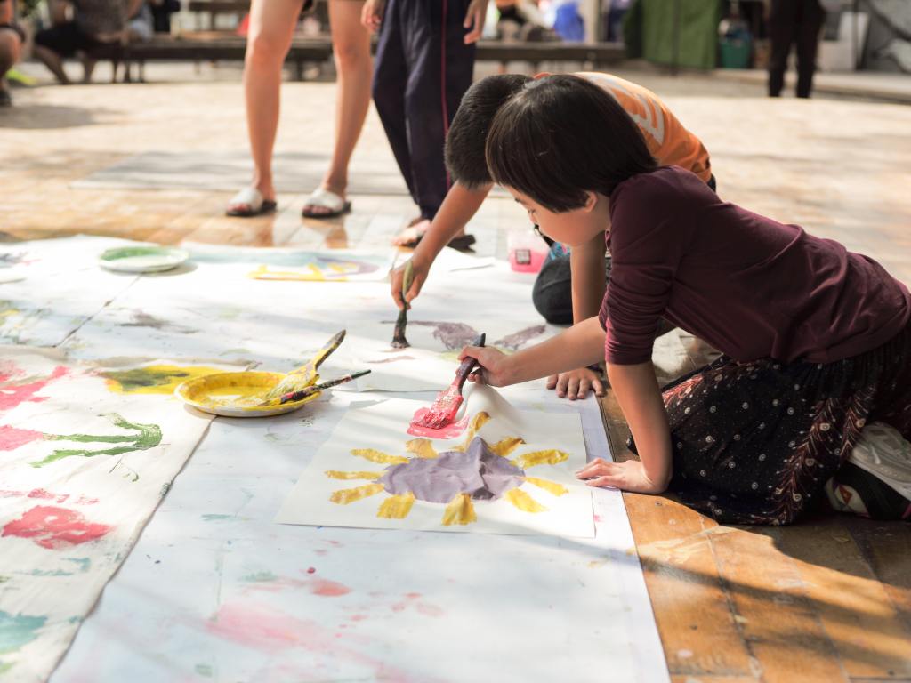 Several children at a homeschool event work together on a large painting on the ground. A plate with paint sits in the middle.