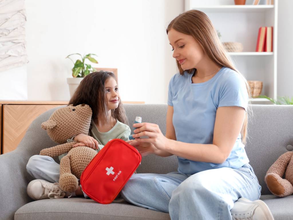 A mom sits on the couch with her daughter. She shows her how to use an inhaler she removed from a red first aid kit.