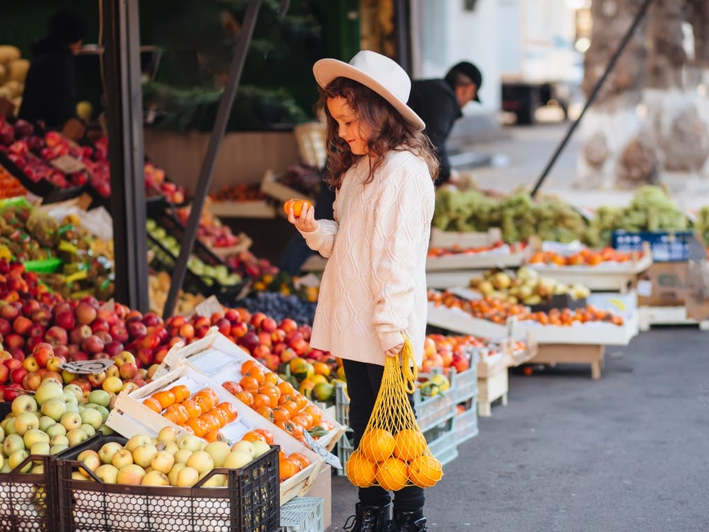 A girl in a cream-colored sweater holds oranges while standing near fruit displays at a farmers’ market.