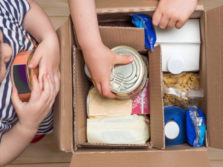 Two young children are adding canned goods to a cardboard box that is full of food items.