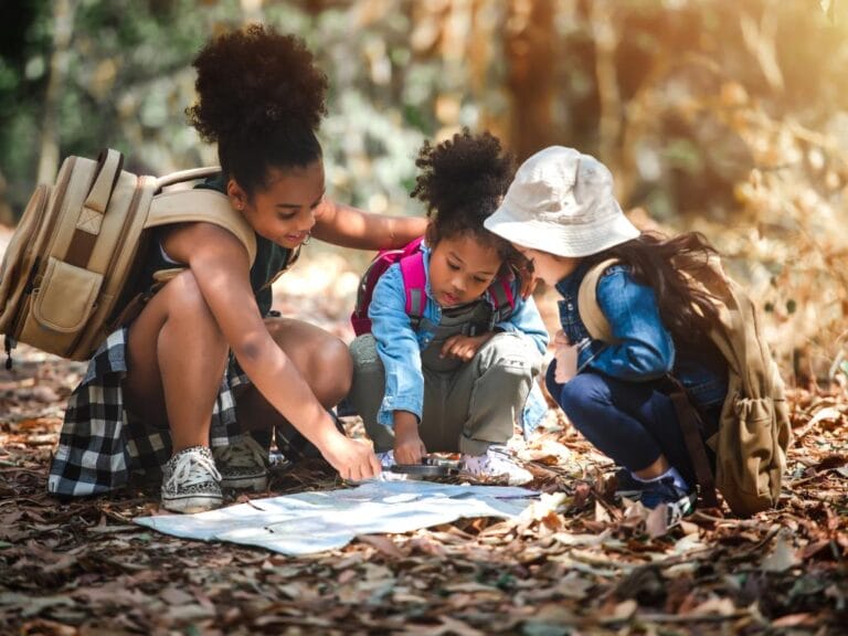 Three small children hold a magnifying glass over a large piece of paper as they huddle in a forest during the day.