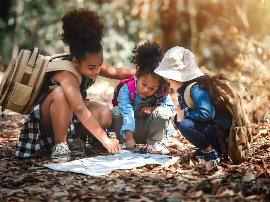Three small children hold a magnifying glass over a large piece of paper as they huddle in a forest during the day.