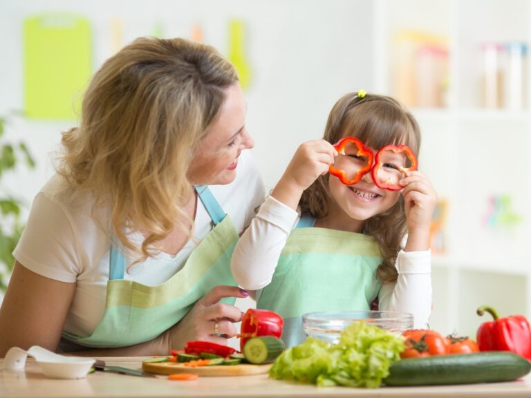 A mother and child sit at a table full of fresh vegetables. The child holds sliced bell pepper rings over her eyes.