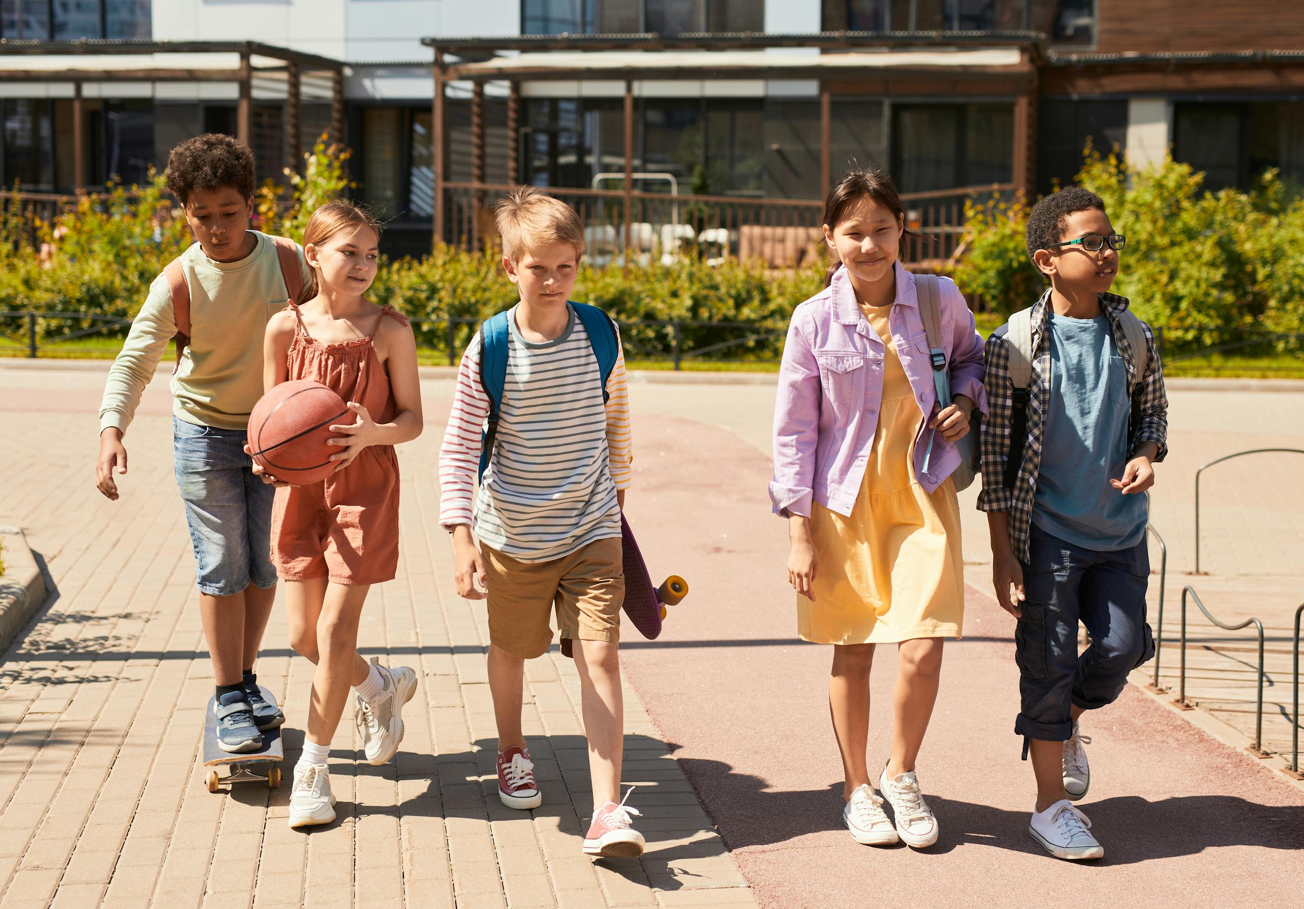 A diverse group of children walking outdoors, carrying backpacks and a basketball, heading to school on a sunny day.