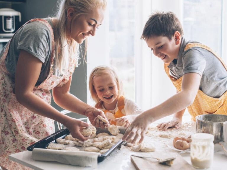 A mother and two kids bake together in a bright kitchen. They shape dough on a counter covered in flour.