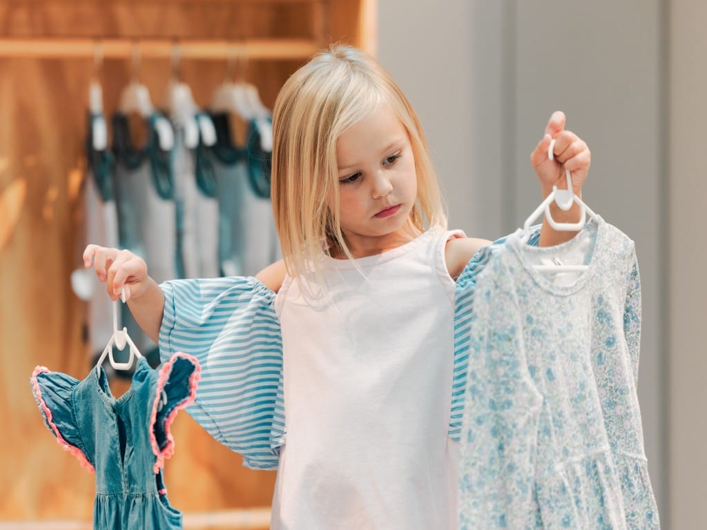 A little blonde girl wearing a white tank top stands in front of her closet. She holds up two different shirts.
