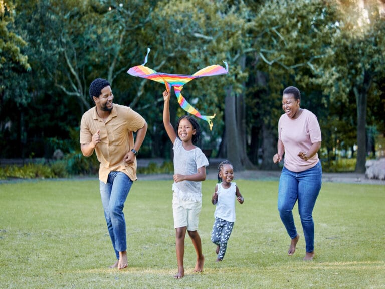 A mother, father, and two little girls run outside in the grass together. One girl holds a rainbow kite above her head.