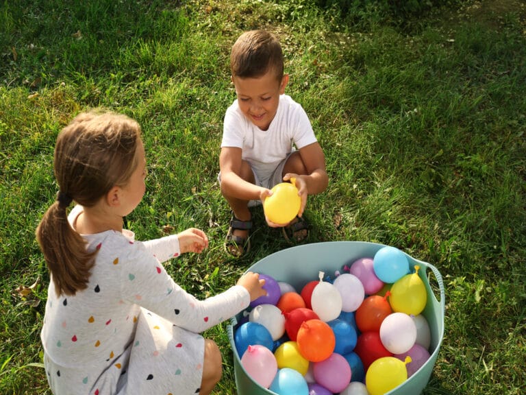 Two kids play on a sunny lawn with colorful water balloons. A boy holds a yellow balloon and a girl reaches into a blue tub.