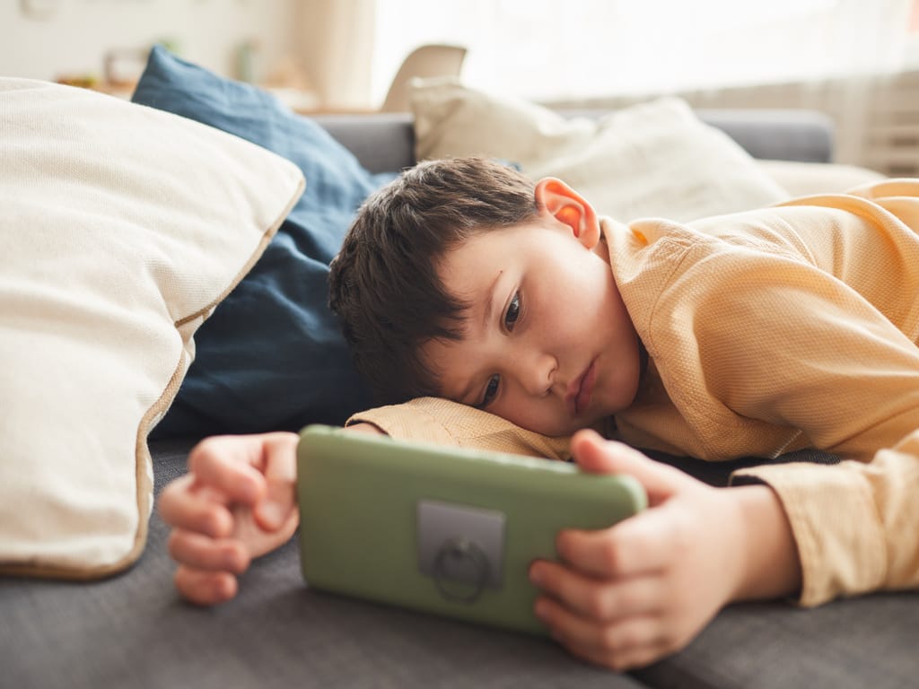 A young boy lies on a bed or couch, holding a green smartphone horizontally with both hands, surrounded by pillows.