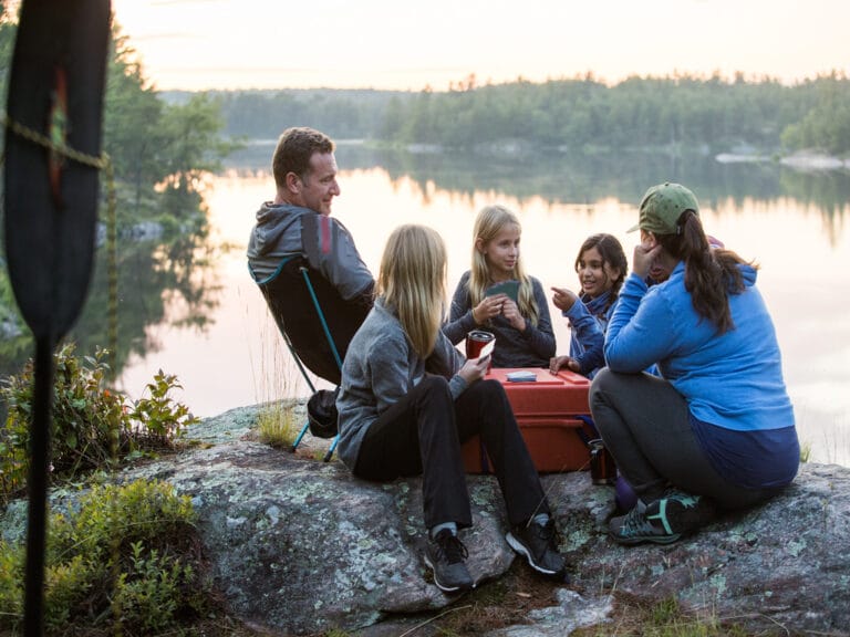 Two adults and three children sit on rocks near a red container, with calm water and distant trees in the background.