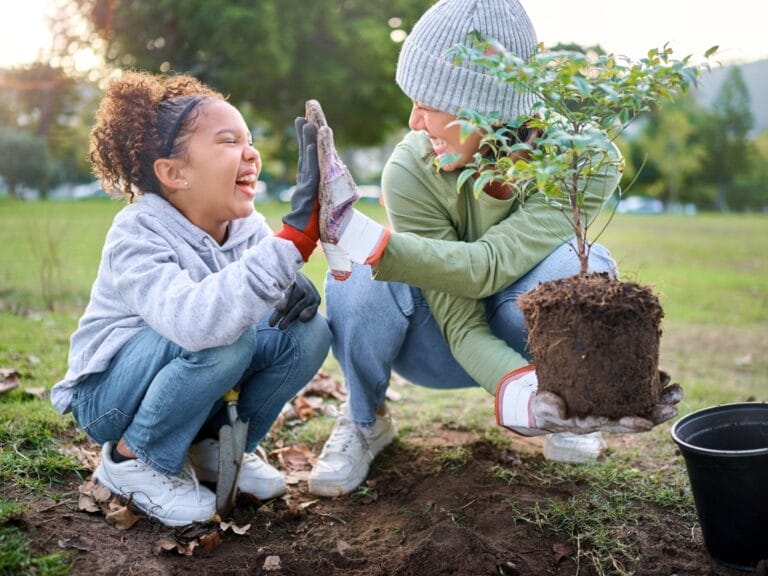 Child and her mom in gloves crouch in a park, laughing and high-fiving while holding a small sapling near a trowel.