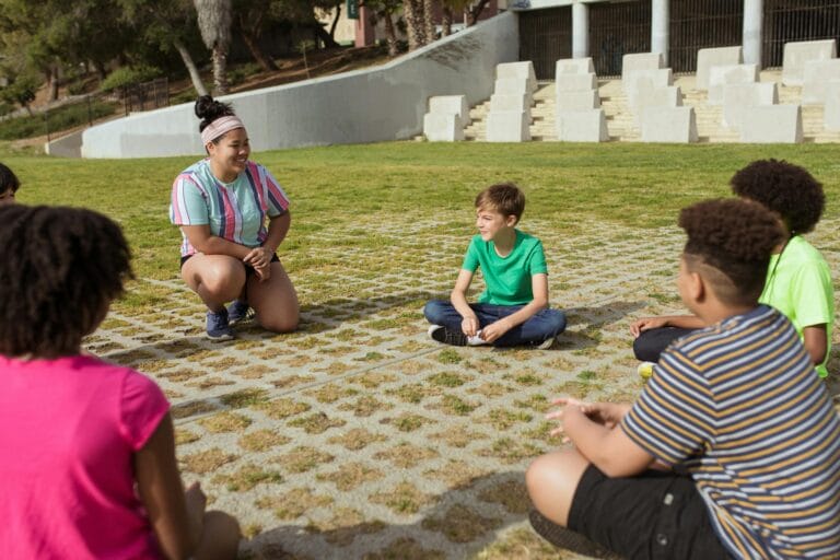 A diverse group of kids sitting in a circle outdoors, engaged in a fun summer camp activity.