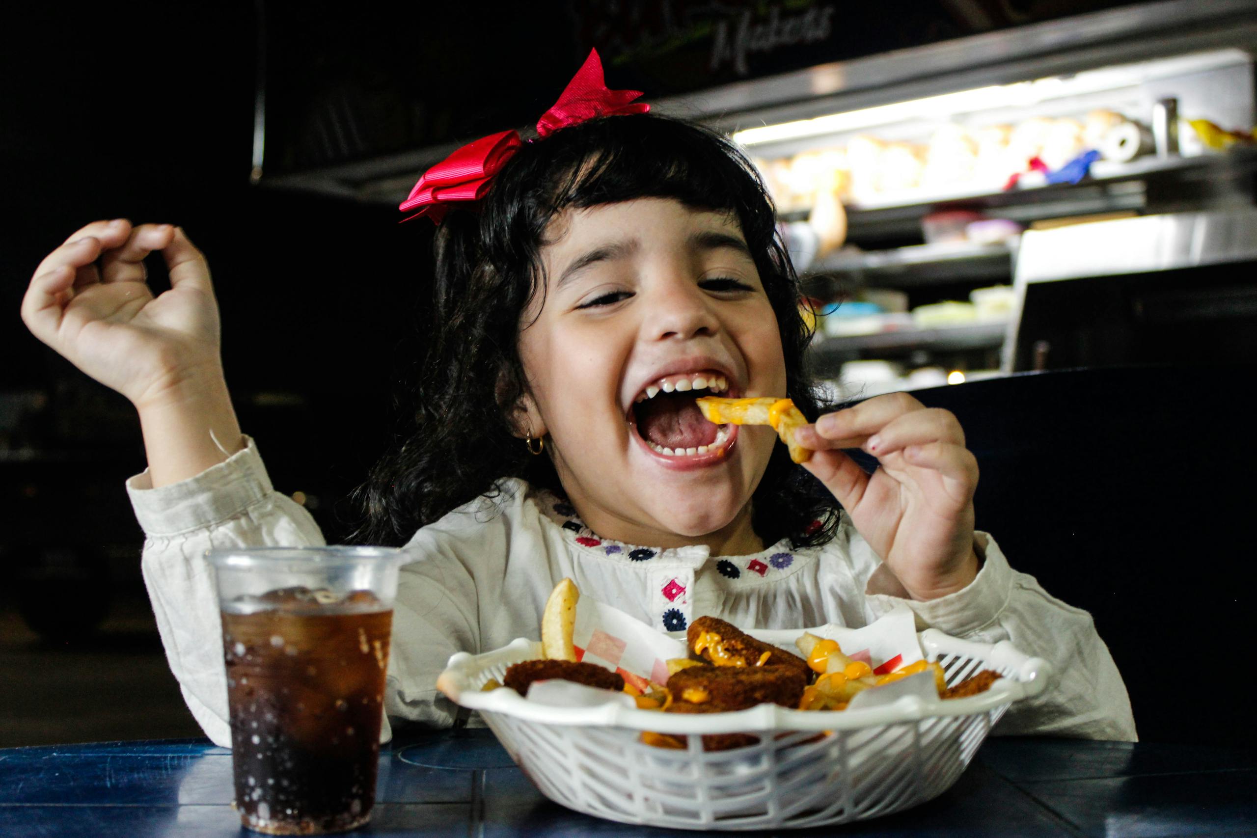 Delighted girl with a red bow eating a basket of fries inside a cafe setting.