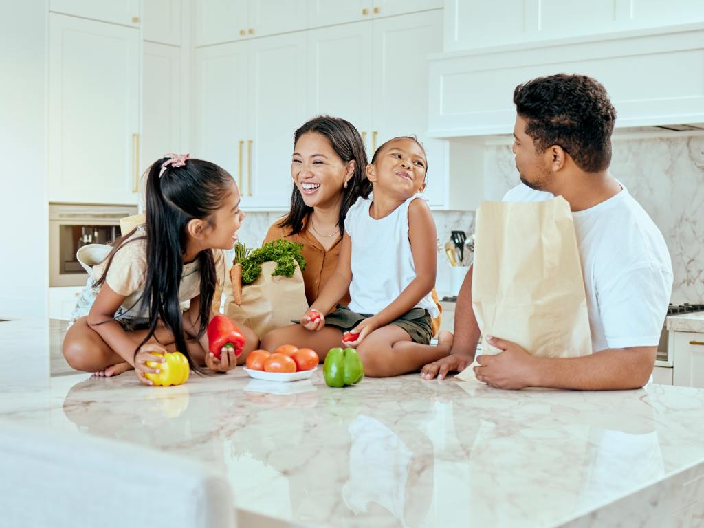 A smiling family sits at a kitchen island sorting fresh vegetables from grocery bags in a bright, modern home.
