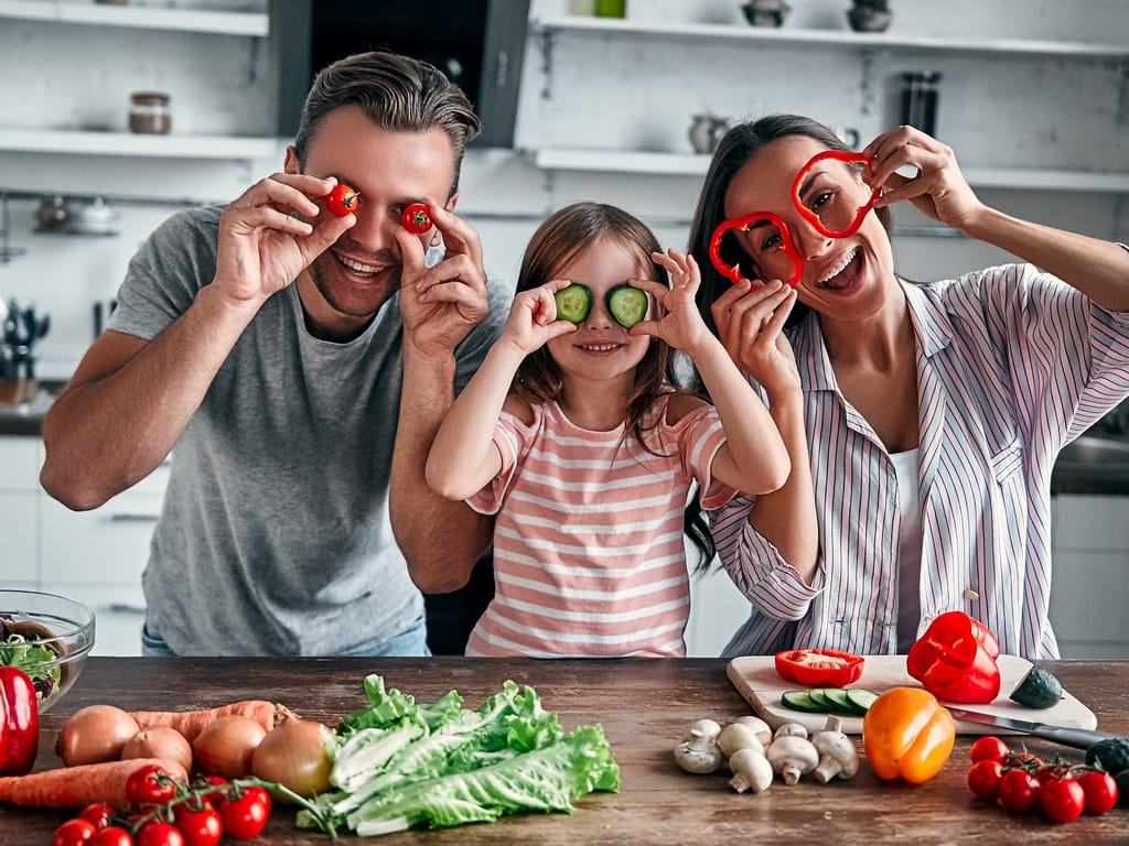 A man, a woman, and a girl in a kitchen hold slices of vegetables up to their eyes. Fresh produce is on the counter.