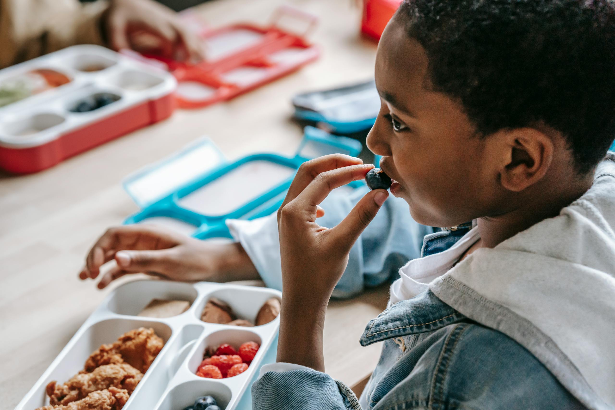 Crop African American child in casual clothes sitting at table with plastic lunch box while having snack