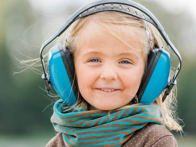 A young child smiles while wearing large blue hearing protection earmuffs and a striped scarf outdoors in soft natural light.