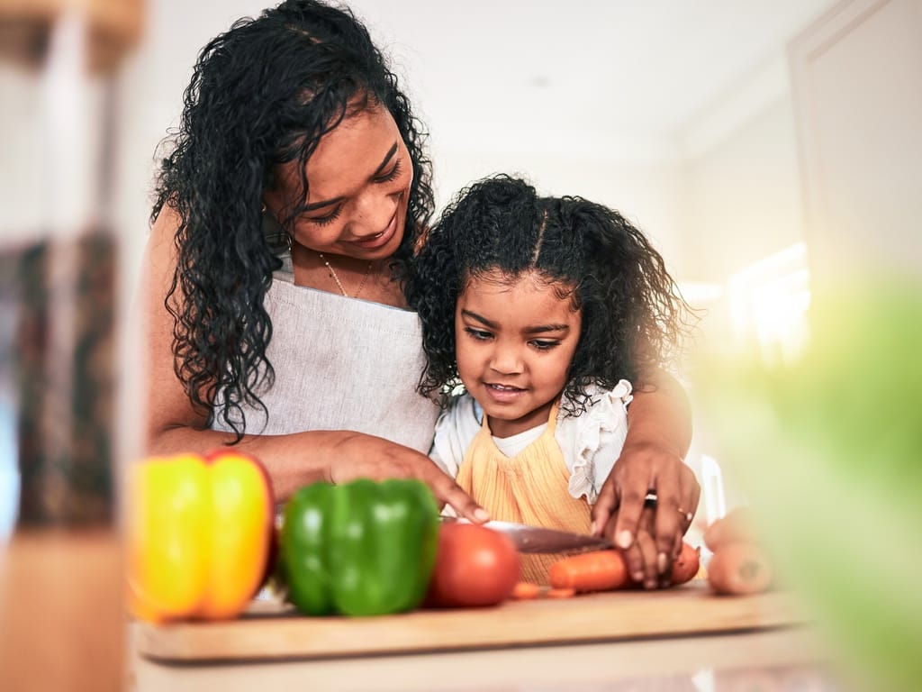 A mother and young girl cut vegetables together at a kitchen counter while cooking and learning at home.