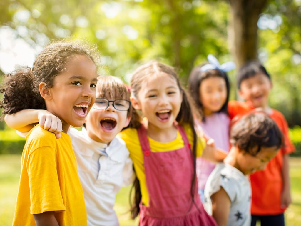 A group of smiling children stand close together in a sunny park, laughing and hugging while playing outdoors among trees.