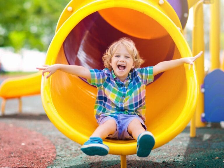 A smiling child wearing a checkered shirt sliding down an outdoor playground slide with arms raised and feet forward.