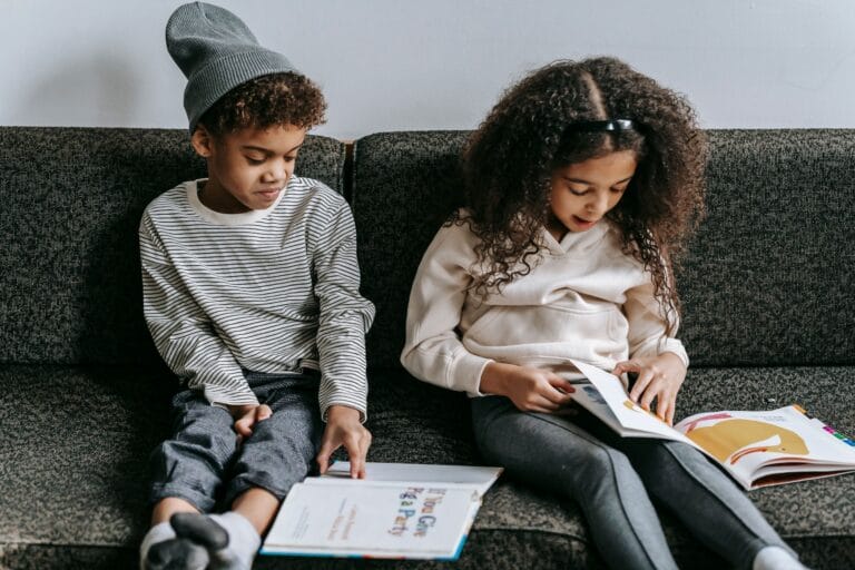 Grimacing stylish African American siblings reading books and sitting together on cozy couch in living room