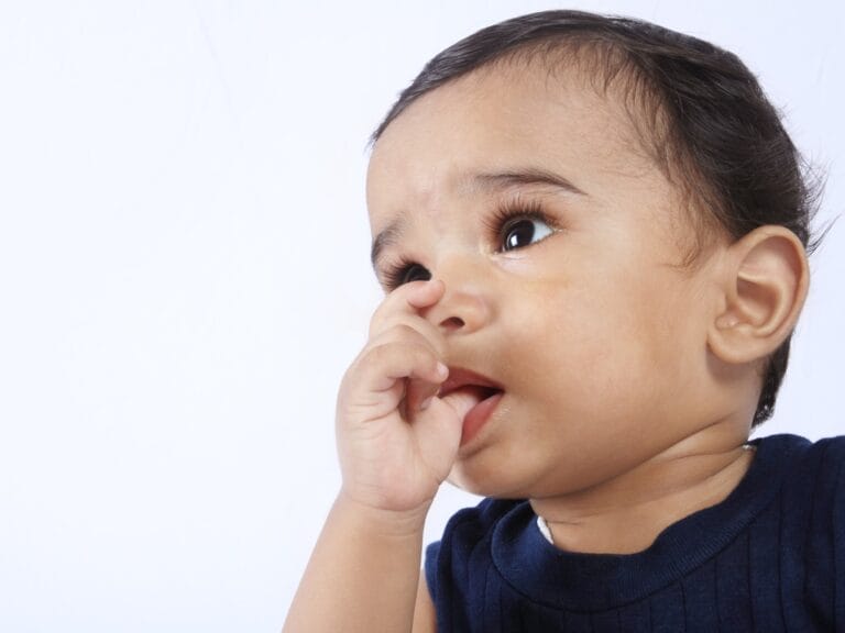 A baby in a navy shirt holding fingers near their mouth while looking upward against a pale white background.