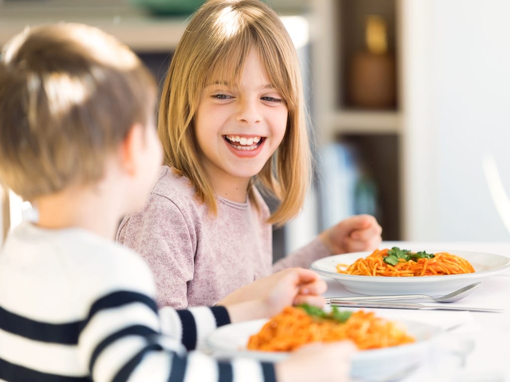 A young smiling girl sits at a table beside a young boy. The bowls on the table have pasta with a red sauce.