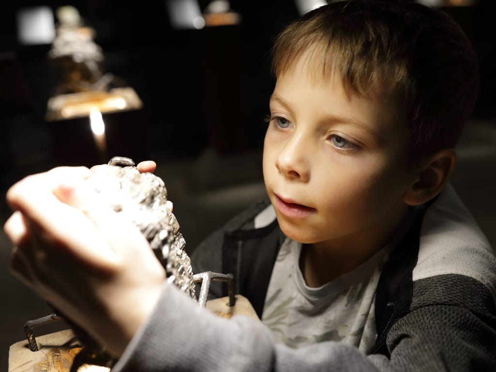a boy closely examining a light-colored mineral specimen under a small lamp in a dark exhibit area while holding it.