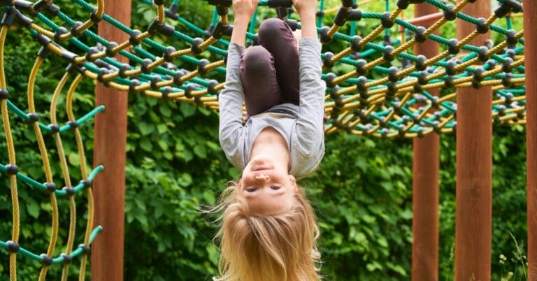 A young child on the playground hangs upside down from a net feature. The playground has wooden support posts.