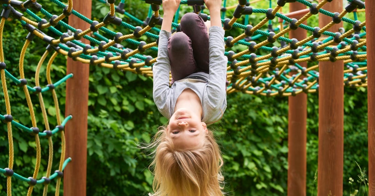 A young child on the playground hangs upside down from a net feature. The playground has wooden support posts.