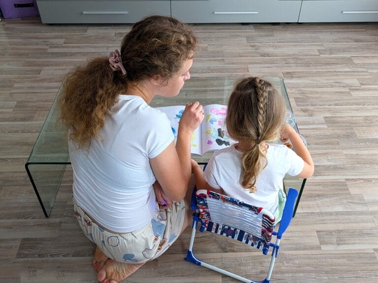 A close-up of an adult and a child sitting at a glass table in a living room, painting in an open sketchbook.
