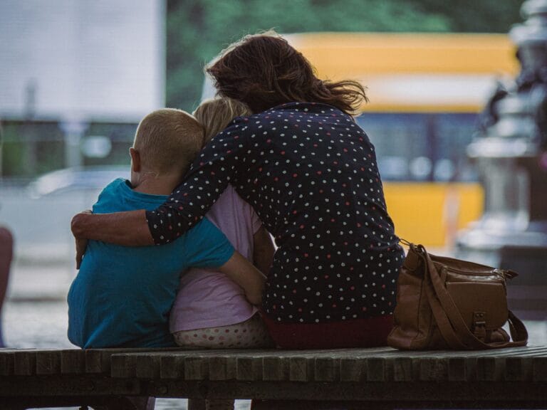 A mother hugging her two children while sitting on a park bench outdoors.