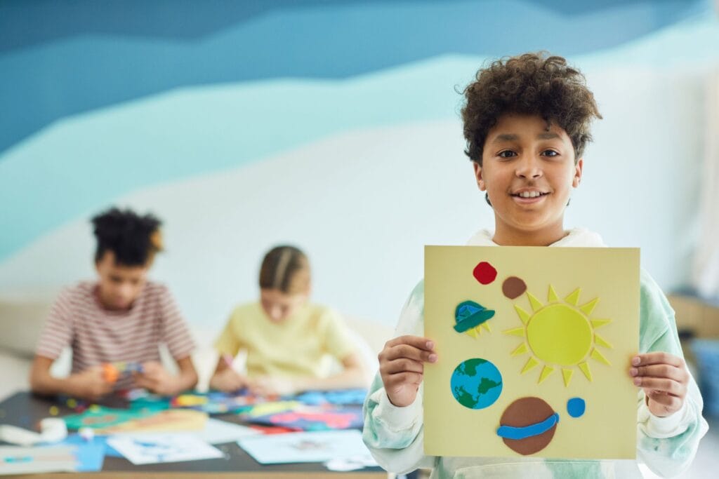 Smiling child displaying a solar system art project in a bright classroom setting.