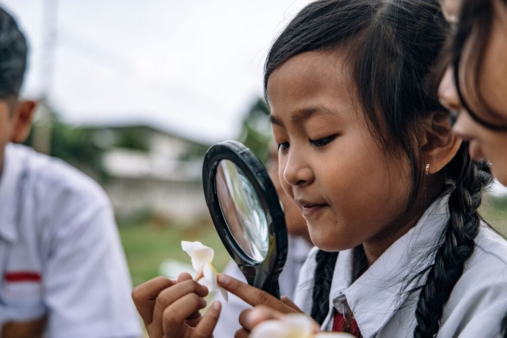Young schoolgirl explores nature using magnifying glass and flower outdoors.