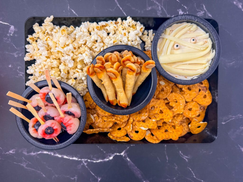 Halloween snack board with finger breadsticks, lychee eyeballs, mummy dip, popcorn, and pretzels.