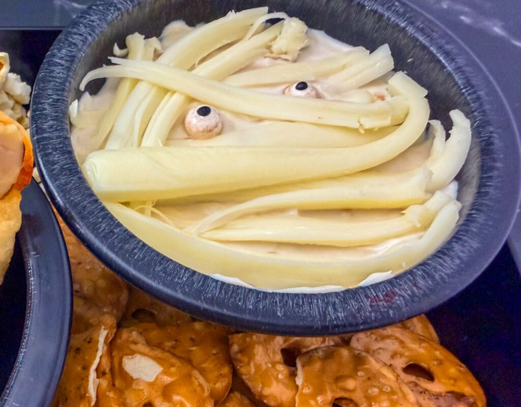 Close-up of Halloween mummy dip with string cheese bandages beside finger breadsticks and pretzels.
