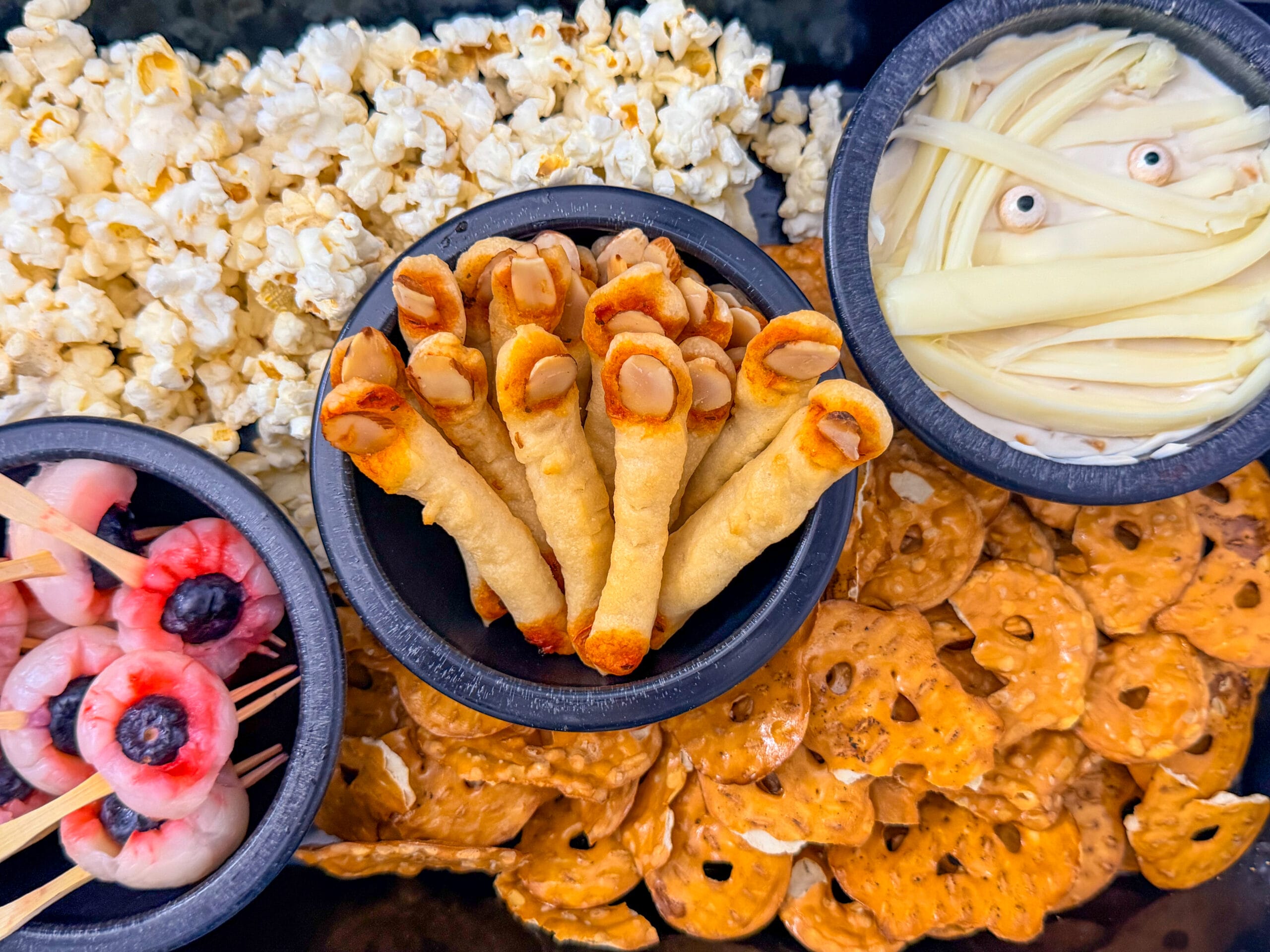 Overhead view of a Halloween party snack platter with lychee eyeballs, finger breadsticks, mummy dip, popcorn, and pretzels.